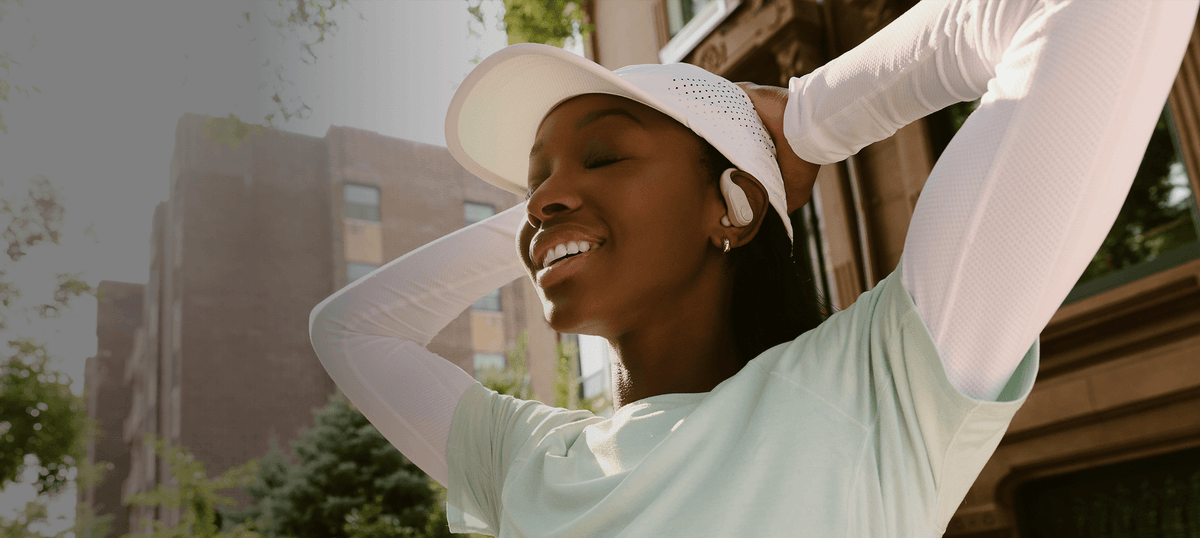 A woman outdoors wearing a white visor, white long-sleeve athletic top, and white wireless earbuds, smiling with her arms raised. Buildings and trees are blurred in the background.