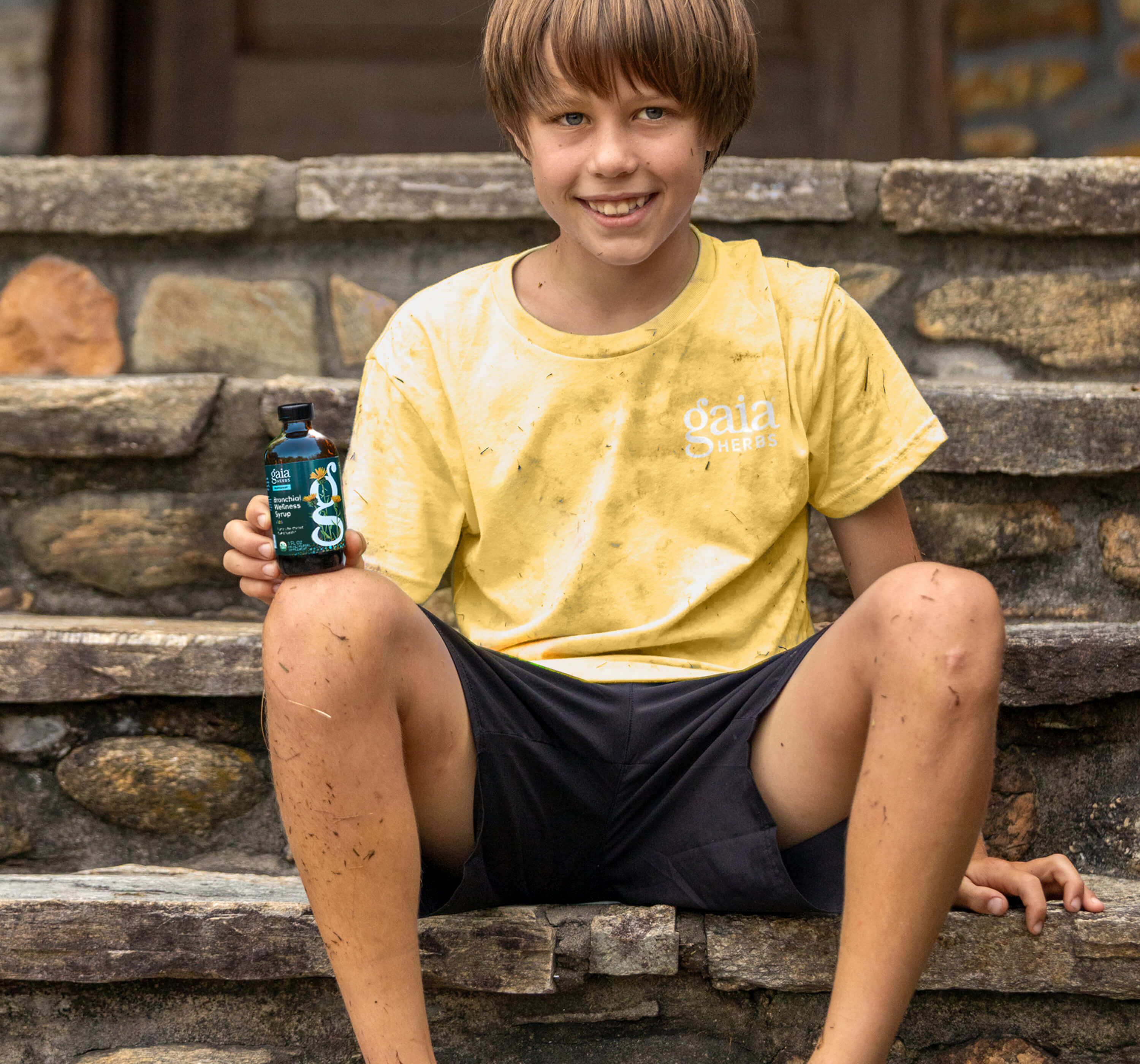 Child holding a bottle of Gaia Herbs Bronchial Kids Syrup on stone steps