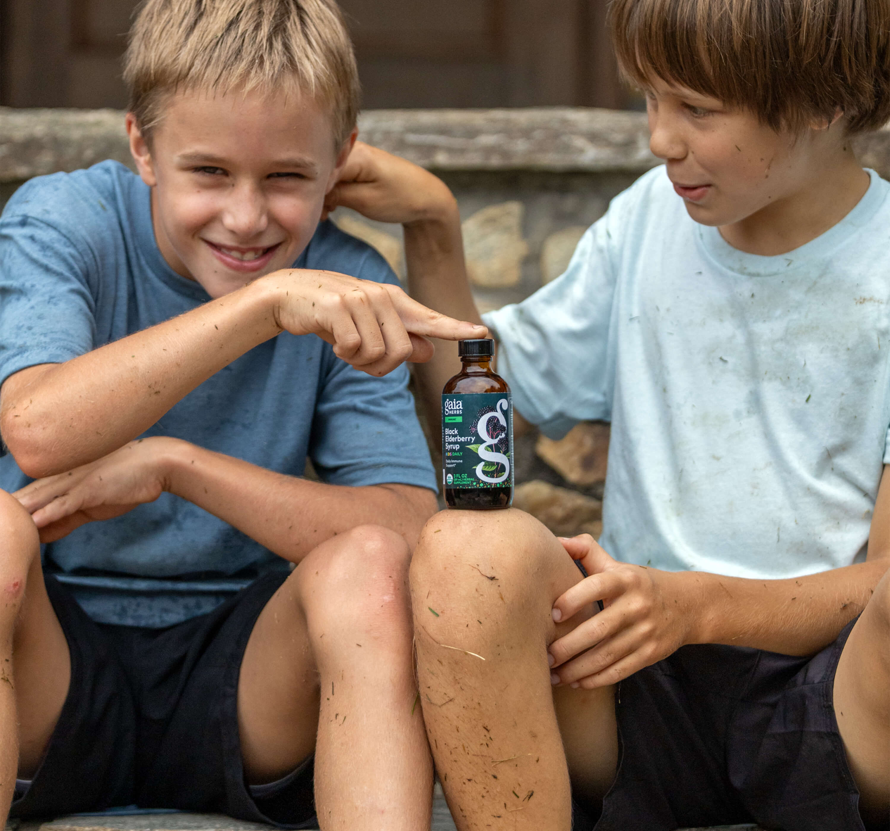 Two children sitting outdoors, one pointing to a bottle of Gaia Herbs black elderberry kids syrup.
