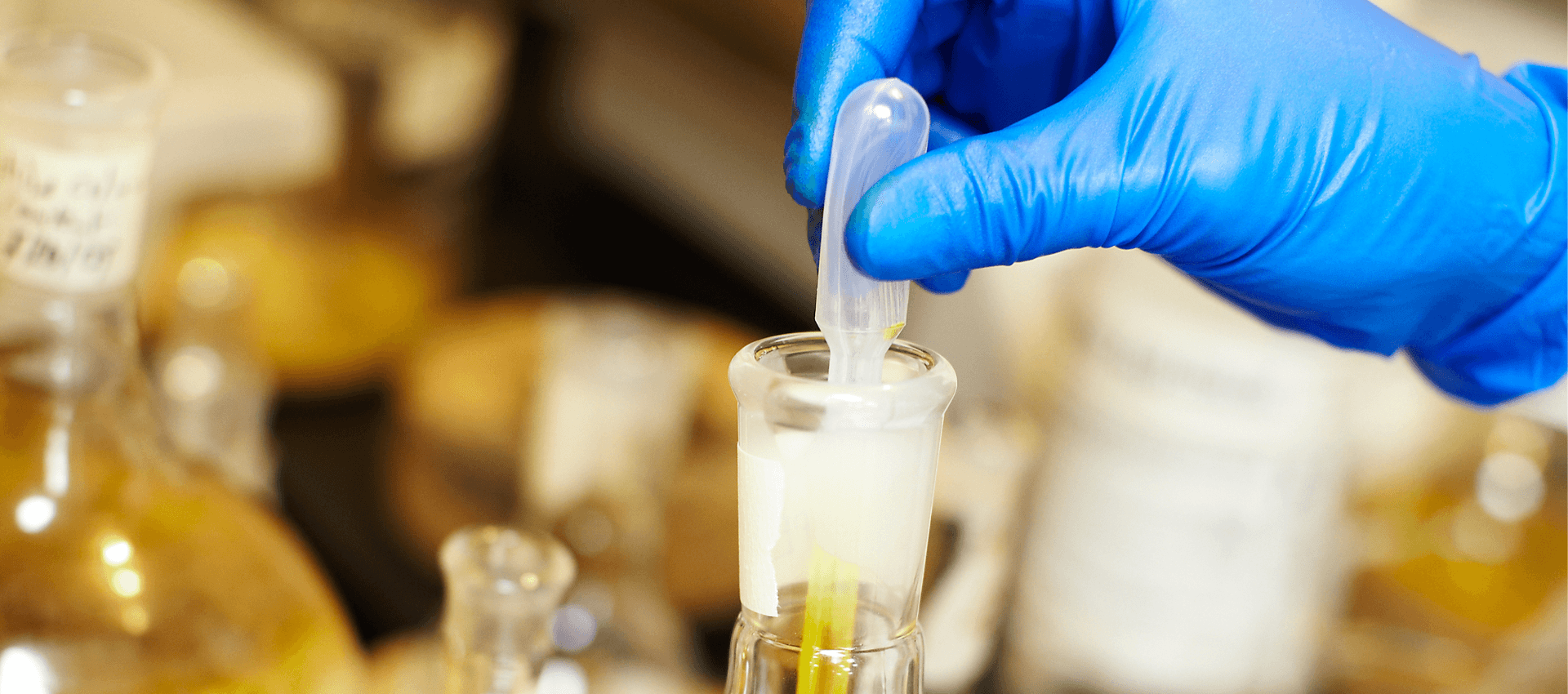 Person wearing blue gloves holding a pipette over a test tube with a blurred laboratory setting in the background.