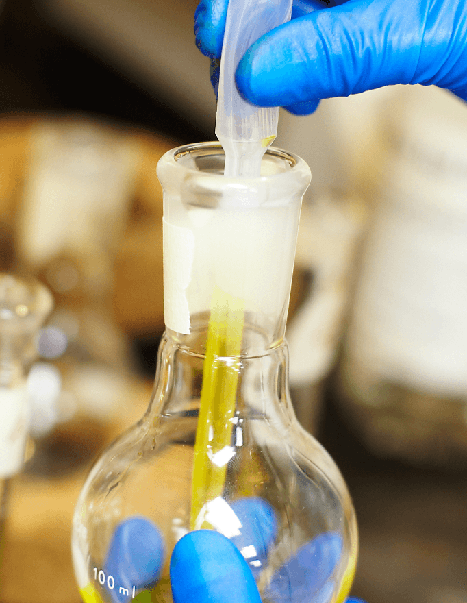 Person wearing blue gloves transferring liquid from a syringe into a glass flask with a blurred background