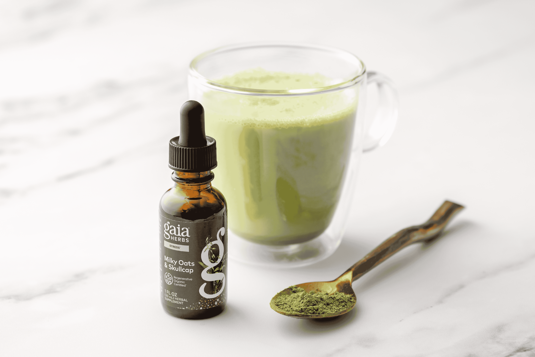 A small amber dropper bottle labeled Gaia Herbs Milky Oats & Skullcap with a glass mug of light green drink, a spoonful of green powder, and a gold spoon on a white marble surface.