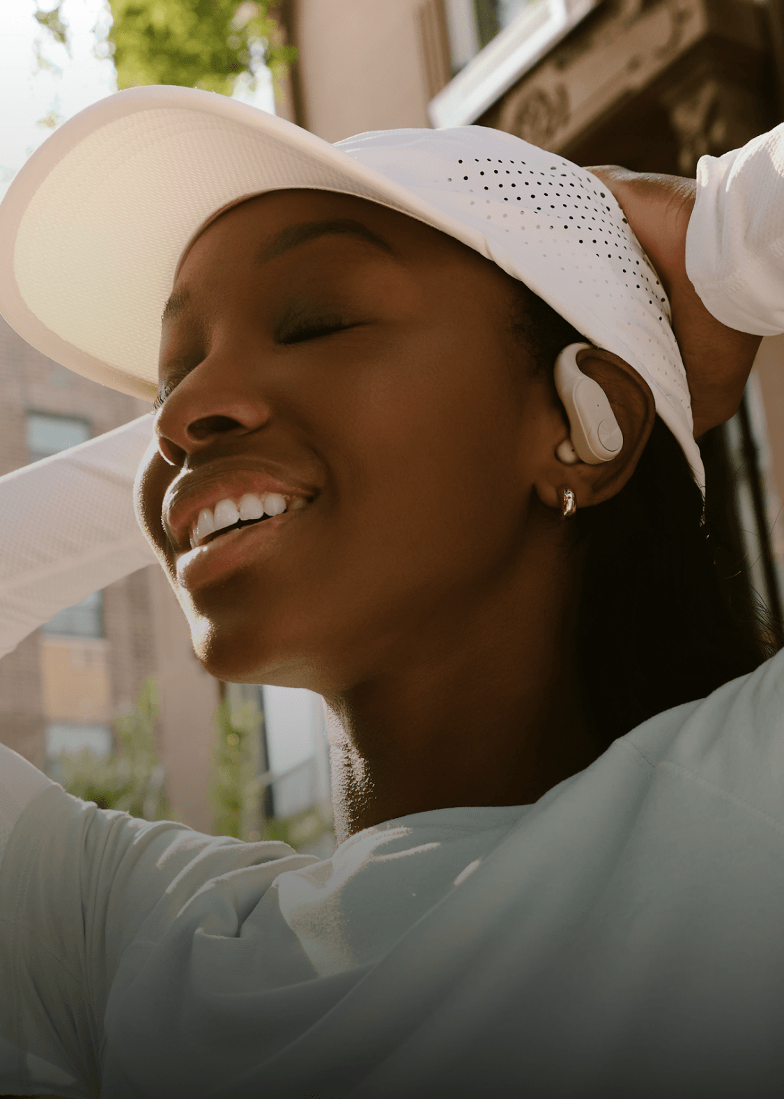 A close-up lifestyle shot of a woman wearing a white baseball cap, a white athletic shirt, and a white wireless earbud, with her eyes closed and a smile on her face.