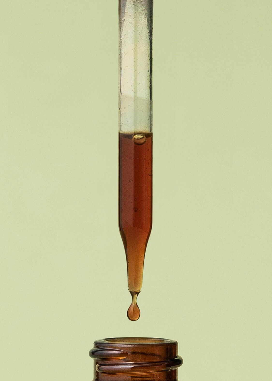 Amber-colored liquid being dripped from a glass dropper into a small glass container on a light green background.