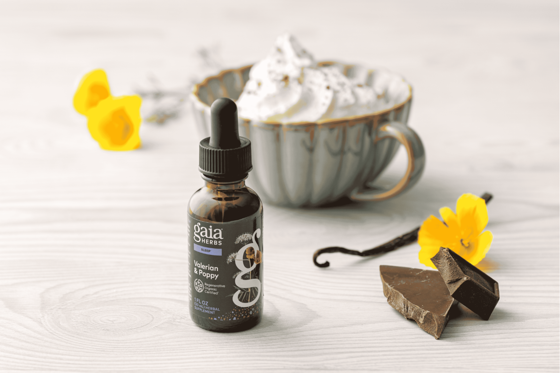 A bottle of Gaia Herbs Valerian & Poppy tincture placed on a light wood surface, styled with a cup of whipped cream, yellow flowers, and chocolate pieces in the background.
